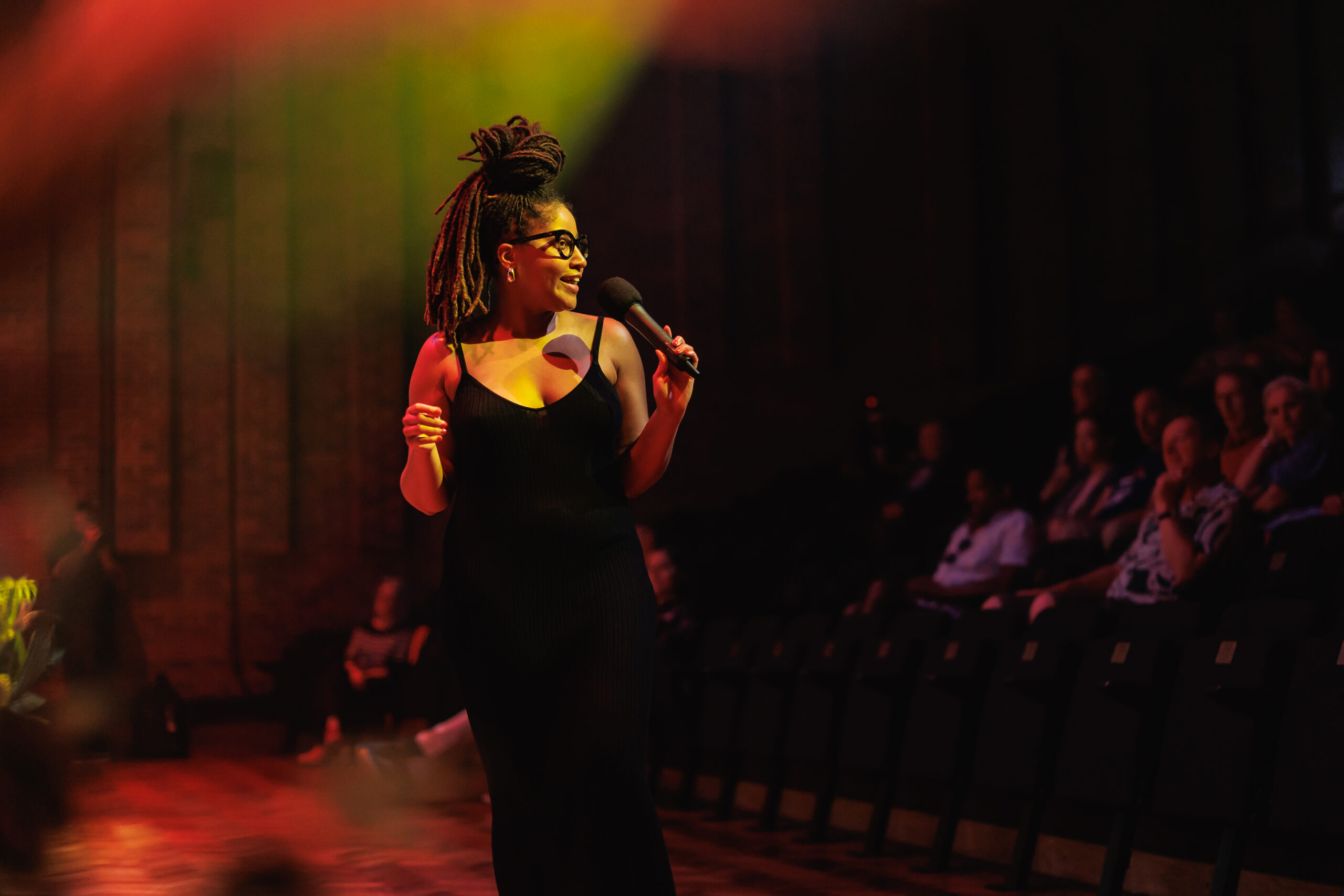 Woman under stage lights in a black dress singing into a microphone and looking towards an audience