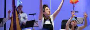 Three female performers with expressive faces and their arms in the air, two wearing night hats. Three music stands and a rainbow umbrella in the background
