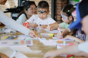 A young boy with Batman facepaint drawing at a craft table with other children.