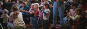 An audience of very young children and their parents seated in an auditorium. SOme of them are standing and clapping