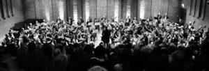 A black and white image of an audience watching a full orchestra on a stage.