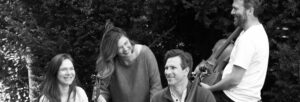 A black and white image of two women and two men, holding string instruments, laughing and sitting on a bench outdoors