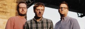 Three men stand outside and gaze at the camera