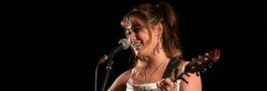 A girl with brown hair is singing into a microphone and playing a guitar.