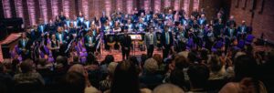 An orchestra standing on stage facing a full audience