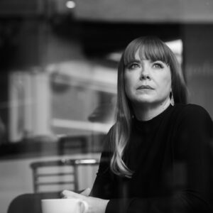 A black and white image of a dark haired woman sitting with a coffee and gazing pensively into the distance