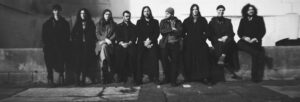 A black and white image of nine people wearing dark clothing sat in a long line along a low wall