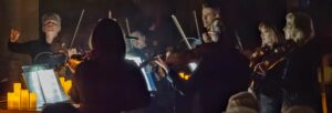 A string quartet standing and playing with an orchestra watching, lit by candlelight