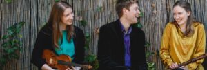 two women and a man, holding musical instruments, sitting outside and laughing