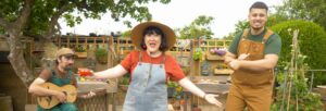 A woman in blue dungarees and a big hat with her arms spread wide and holding a small tomato with eyes, with a man in brown dungarees and a seated man playing the guitar.
