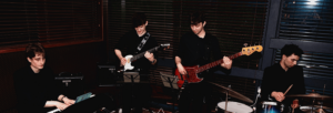 A keys player, guitarist, bass player and drummer stand in a dimly lit room playing music.