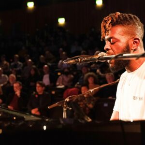 A black man in a white tshirt singing into a microphone and playing the piano to an audience