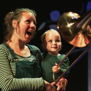 A blonde woman holding a small blonde child and looking in surprise at a trombone