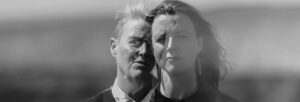 A black and white image of two woman standing in front of water. Their images are layered over each other.