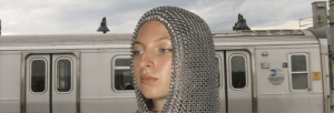 A girl wearing a chainmail hood stands in front of a train.
