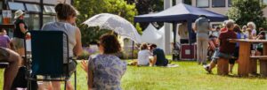 People sitting and talking outdoors, sat on the grass and at a picnic table with a small stage under a marquee in the background