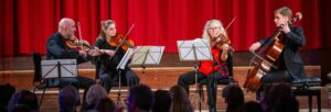 Four musicians, two men and two women, playing stringed instruments on the stage in front of an audience with a red curtain backdrop