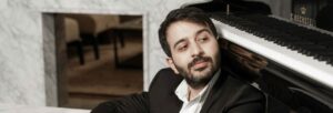 A man with black hair and a black beard a moustache sits with his back to a piano.