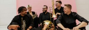 A group of four young men and one young woman holding brass instruments and laughing together
