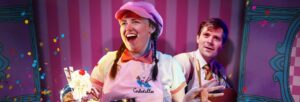 A smiling woman in a pink flat cap with an ice cream in front of her. A man looking amused and surprised stands behind her.