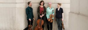 A man and three women holding string instruments standing in front of a taupe wall