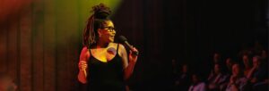 Woman in a black dress and glasses holding a microphone and smiling at the audience at Turner Sims Southampton