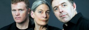 Two men and a woman, all wearing black, gazing at the camera with serious faces, against a grey background