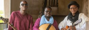 Three men stand together, with two of them holding guitars and one of them holding a wooden drum.