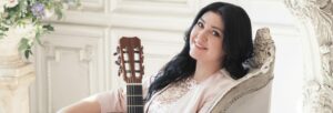A woman with black hair sits in a white chair while holding a guitar.