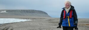 A white haired lady in heavy-duty outdoor clothing standing in a bare landscape next to rocks