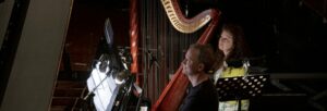 A female harpist with long black hair and a male pianist with short, brown hair both sit at their instruments.