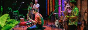 Man in dungarees and a colourful red shirt playing the keyboard and singing with rainbow lights and a band in colourful dungarees and shirts in the background