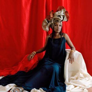 A black woman wearing a blue dress and a large headdress, sitting on a chair draped in white fabric before a red fabric backdrop