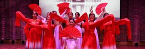 A group of women wearing traditional red and pink Chinese clothes stand together holding paper fans.