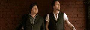 Two men stand together, one holding a fiddle and the other holding an accordion.
