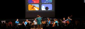 A group of classical musicians wearing blue shirts perform in front of an audience. A screen with colourful images is behind the stage.