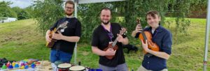 Three smiling men, each playing a different stringed instrument, stood behind a table covered in various instruments.