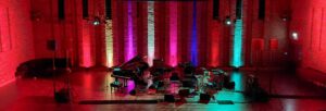 a grand piano and other instruments on stage under jewel coloured lighting in an empty auditorium