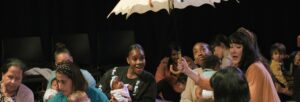 A group of various mothers with their babies, sitting on mats on the floor whilst a woman holding a parasol sings