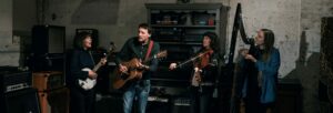 Three women and one man playing instruments, standing in a dimly lit room surrounded by amps and speakers