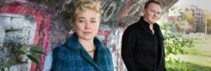 A blonde woman in a blue check coat and a man in a black coat stood under a graffitied bridge