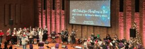 A group of musicians are performing in Turner Sims, dressed in Christmas hats and other festive wear. A choir stands on the left of the stage, and a small orchestra is seated on the right. In the middle stands a female guitarist, male saxophonist and male bassist.