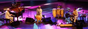 Three black men in white African dress playing piano and percussion on a purple lit stage