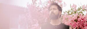A man with a beard wearing a black tshirt standing in front of a pink blossom tree