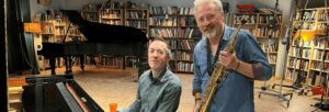 A man holding a saxophone standing next to a man seated at a piano with shelves full of books in the background