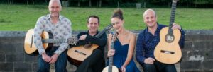 Three men and one woman, all holding guitars, sitting on a wooden bench in front of a grey brick wall with grass behind them
