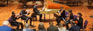 A group of young people playing classical instruments, including an antique harpsichord, on a stage with a brick wall behind them