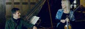 A dark haired man sitting at a grand piano smiling at a blonde woman holding a violin