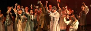 A large group of men and women on stage wearing Dickensian dress, raising their glasses to each other