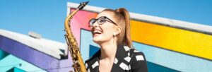 A laughing woman with a high ponytail and glasses holding a saxophone against a bright blue sky